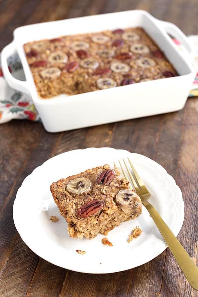Slice of Banana Bread Baked Oatmeal on white plate with fork in front of white baking dish